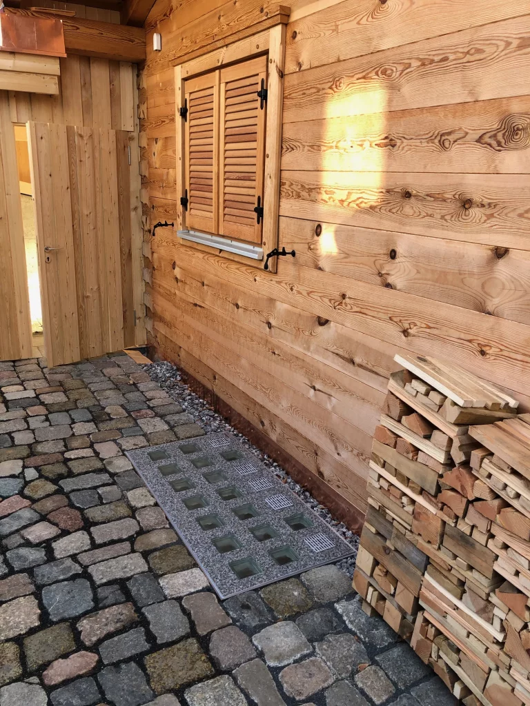 Hochwertige wasserdichte Lichtschachtabdeckung an einem Holzhaus mit Kopfsteinpflaster