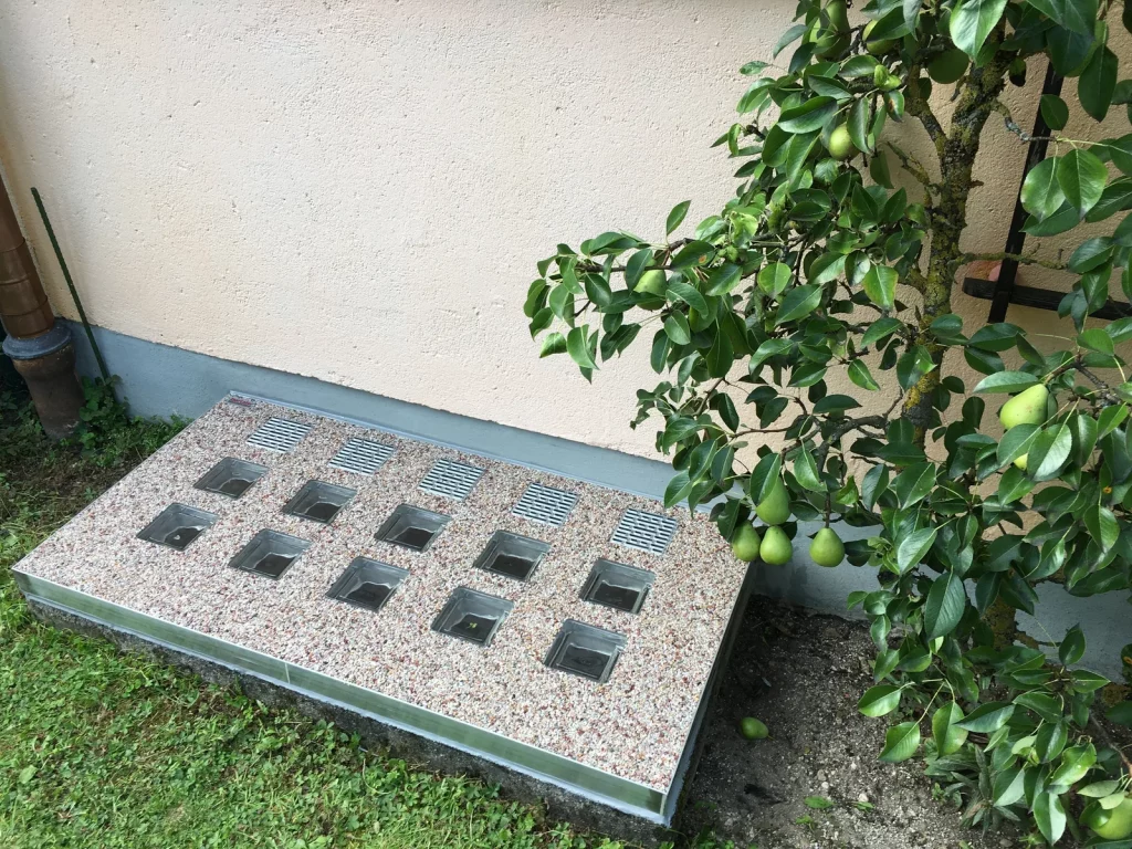 Maßgefertigte wasserdichte Lichtschachtabdeckung mit Steinteppich und Glasbausteinen neben einem Birnbaum